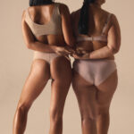Rear view of two female models in soft neutral underwear holding hands in gentle connection. Concept of inclusive fashion storytelling, intimate branding, body-positive visuals, diversity promotion.