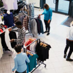 Top view of diverse customers checking stylish clothes, shopping for fashionable merchandise and accessories in modern boutique. Clients analyzing hangers with new fashion collection